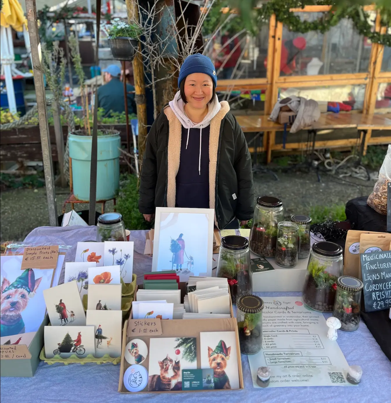 Amy stands behind her market stall, smiling, with watercolour cards, prints and glass terrariums laid out on the table under evergreen garlands.