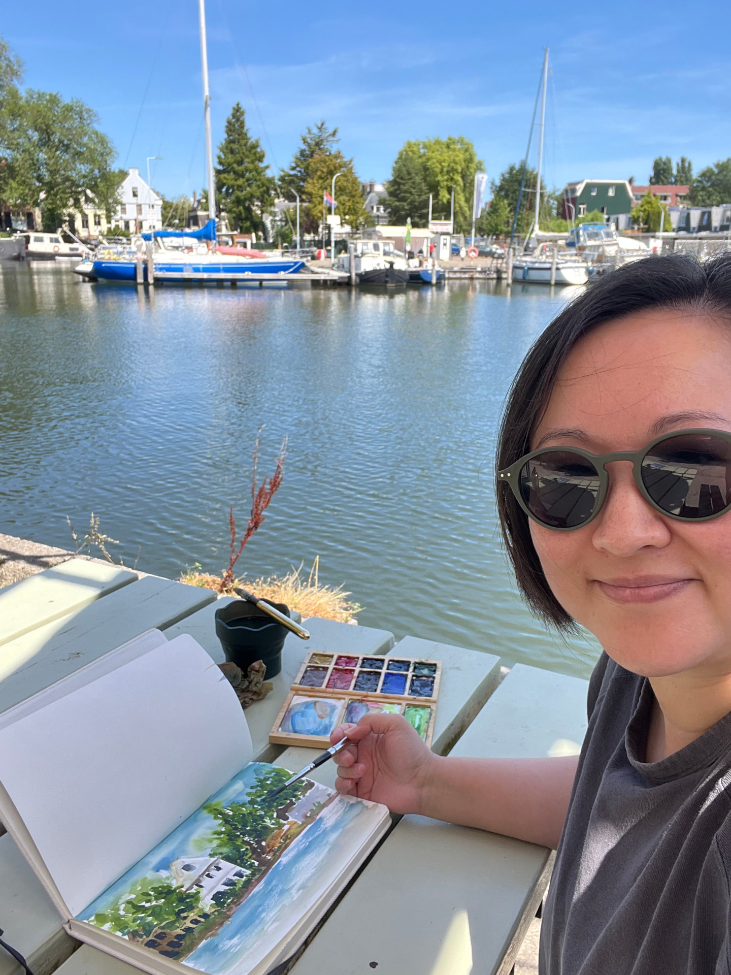 Amy painting a watercolour landscape outdoors by the water, with her sketchbook and paint palette on the table