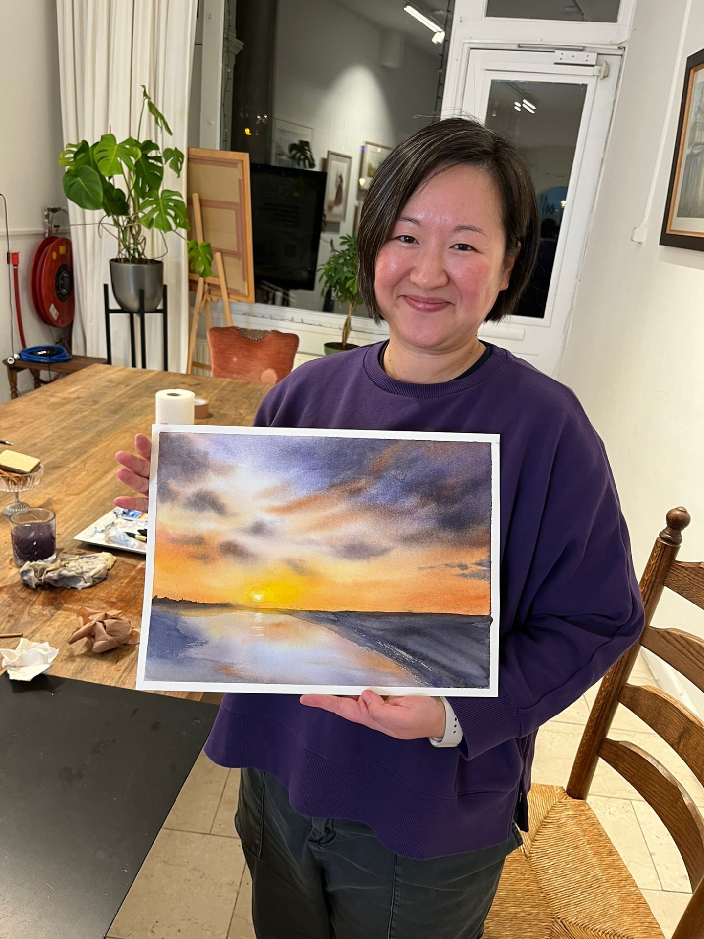 Amy holding a watercolour painting of a sunset over the water in her studio